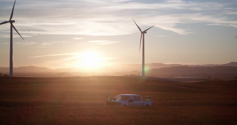 A wind farm engineer inspects spinning turbines from a rural field next to a pickup truck during a beautiful golden hour sunset