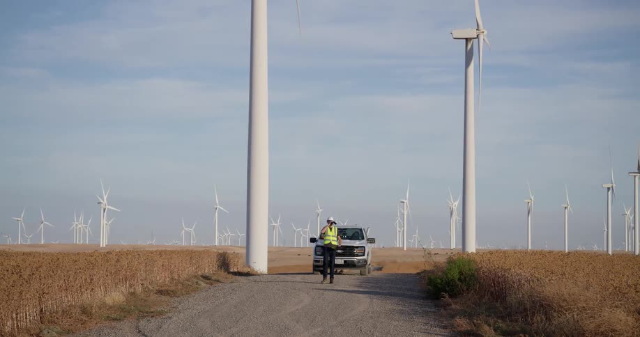 A male engineer walks away from his truck towards the camera on a dirt road while inspecting a vast wind farm in a rural field