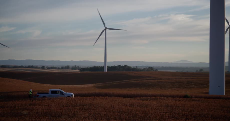 An engineer surveys a vast wind farm of spinning turbines across golden rolling hills from their truck during a beautiful sunset