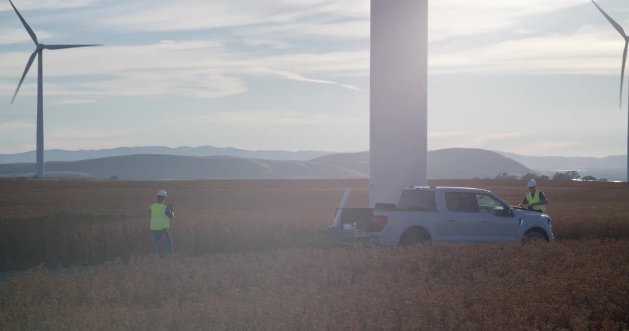 Two engineers in safety gear perform a site survey at a vast wind turbine farm during a golden sunset