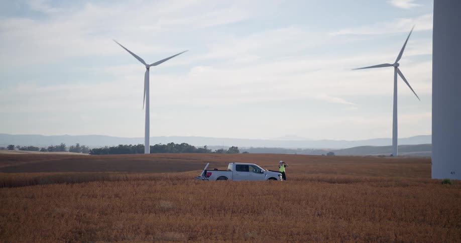 A slow pan reveals a wind turbine technician working next to his truck in a vast golden field surrounded by spinning windmills