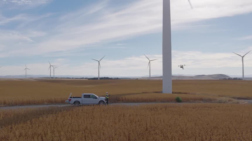 A technician launches an inspection drone near a massive wind turbine, with a vast wind farm stretching across the rural landscape