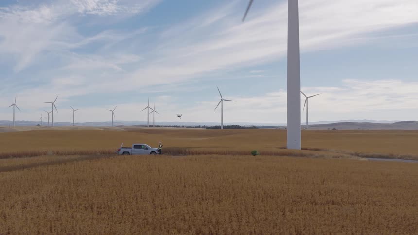 A technician operates a drone to inspect a large wind turbine at a wind farm situated in a vast golden field