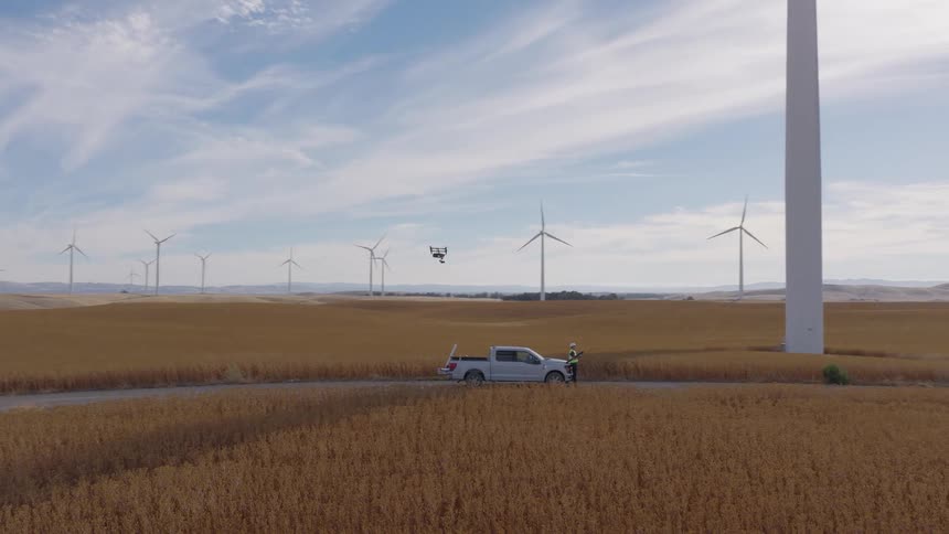 Aerial view of a technician operating a drone for wind turbine inspection on a vast wind farm surrounded by golden fields
