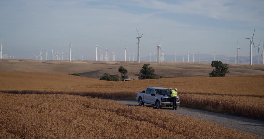 Engineers conduct an inspection at a sprawling wind farm, standing by their truck in a vast golden field under a clear sky