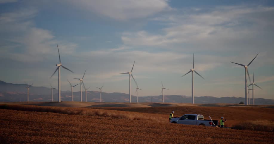 A panoramic view reveals a sprawling wind farm on rolling golden hills, with engineers working near a turbine at sunset