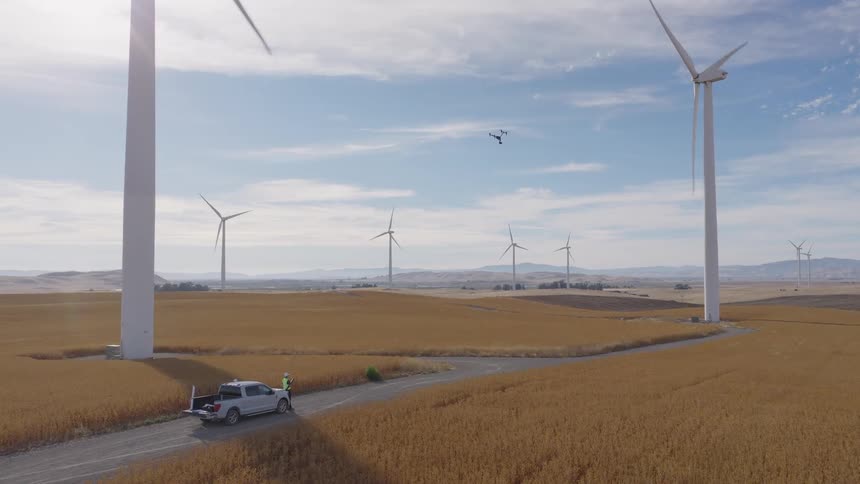 Technicians launch a drone to conduct an inspection of a large wind turbine on a sunny day at a wind farm