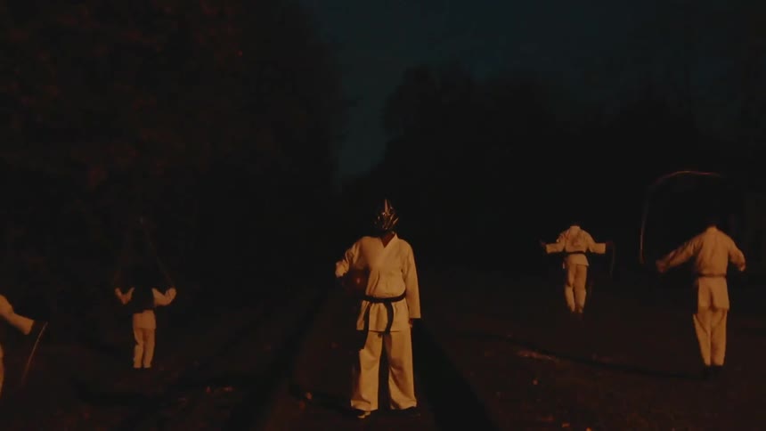 A surreal and mysterious scene of a masked martial artist standing on train tracks at night surrounded by headless figures jumping rope