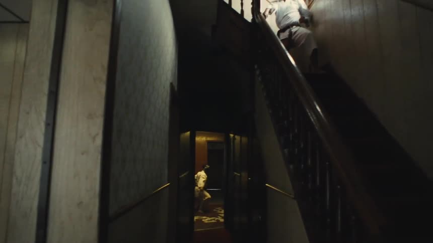 Two masked martial artists train in a dimly lit, suspenseful house, one on the stairs and one in the hallway below