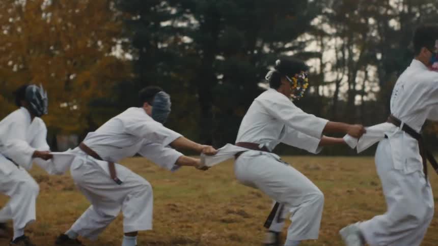 Masked martial artists in white uniforms engage in a ritualistic tug-of-war in a grassy field