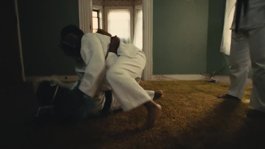 A martial artist in a white gi and black belt throws their opponent to the ground in slow motion during an indoor practice session