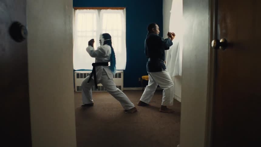 Seen through a doorway, a man and woman in martial arts attire practice a synchronized kata in slow motion inside a sunlit room
