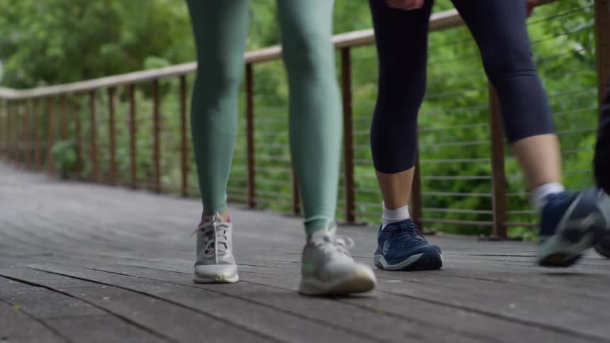 Low-angle slow-motion shot of three women's legs and feet walking along a wooden boardwalk in a park