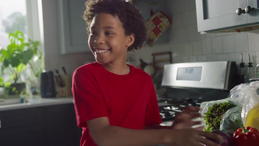 A happy young boy playfully examines a red onion and bell pepper while helping unpack groceries in the kitchen