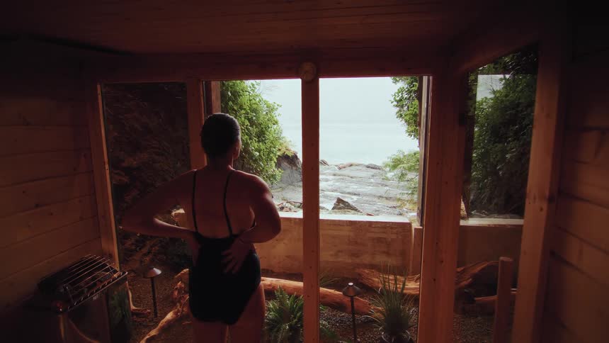 A woman in a swimsuit looks out from a rustic wooden sauna onto the rocky ocean coastline on an overcast day