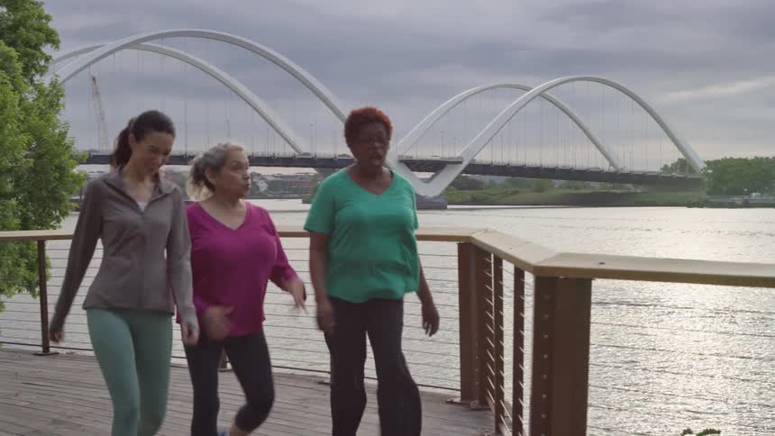 Three women of different ages and ethnicities enjoy a walk and conversation along a riverside boardwalk with a modern bridge in the background