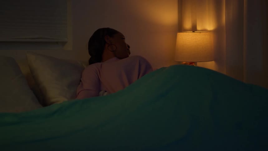A woman gets into bed in her dimly lit bedroom, turns off the lamp with a remote, and settles in to sleep for the night