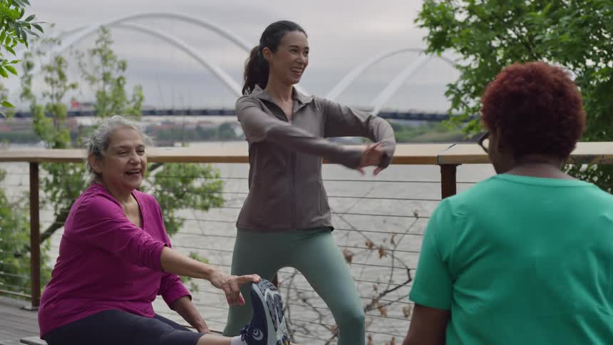 A smiling fitness instructor leads two senior women in a joyful stretching routine on a wooden deck overlooking a city river