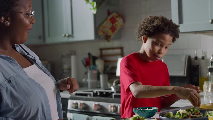 A young African American boy helps his mother make tacos for dinner, theatrically adding the final toppings and giving an 'OK' sign of approval