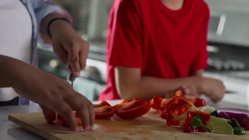 Close-up of hands slicing a red bell pepper on a cutting board as a smiling boy watches and tastes a fresh piece in the kitchen
