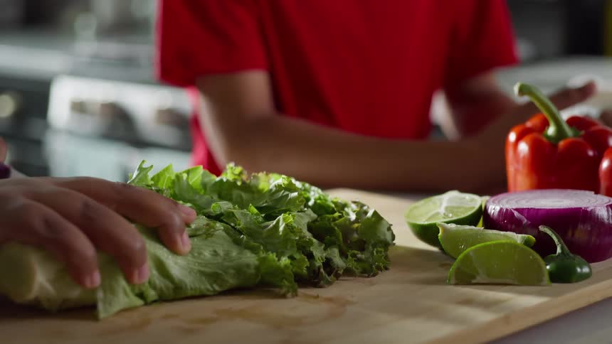 Slow-motion close-up of hands slicing fresh lettuce on a wooden cutting board while a child watches in the background