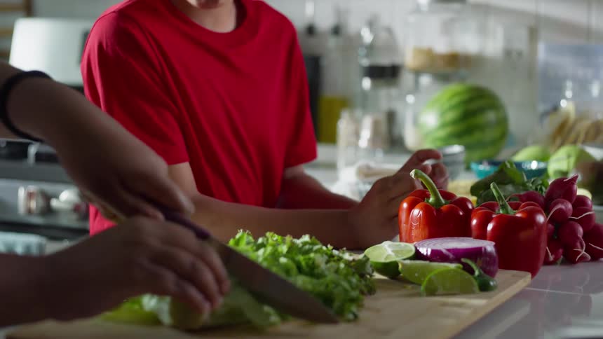 A smiling young boy watches a person chop fresh lettuce on a cutting board in a sunlit kitchen