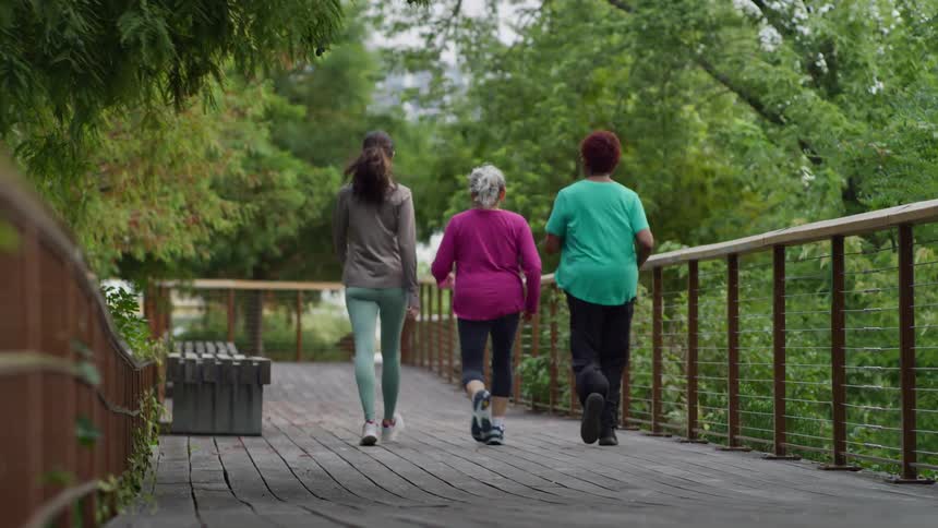 A slow-motion, rear-view shot captures a young woman and two senior women walking together on a wooden boardwalk in a serene park
