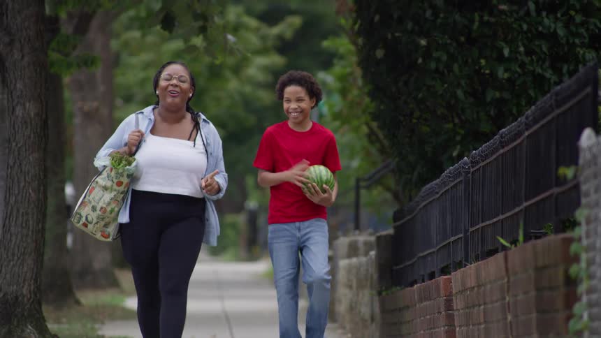 A happy mother and son laugh and talk as they walk home from the grocery store carrying a watermelon and a bag of produce down a leafy sidewalk