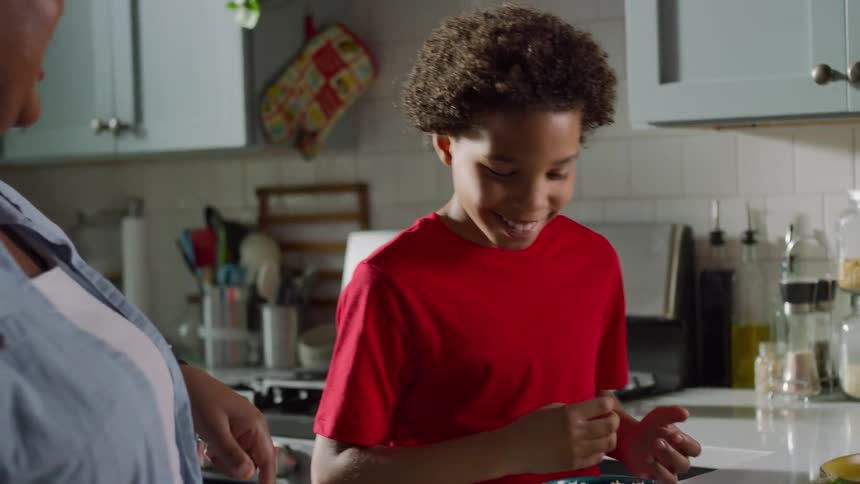 A happy mother and son laugh together in slow motion as they sprinkle cheese on fresh tacos in their bright kitchen