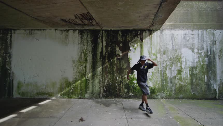 A young man shows off his impressive hip-hop footwork in slow motion under a gritty urban overpass