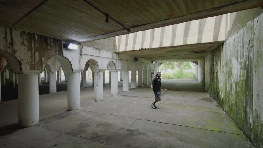 A man in a hat skillfully juggles a soccer ball in slow motion inside a gritty, abandoned concrete underpass with repeating columns
