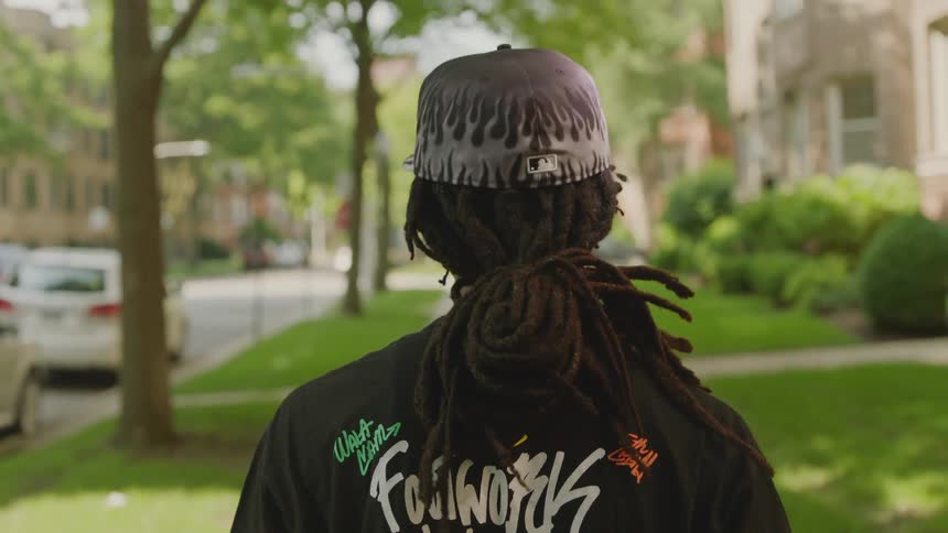 Over-the-shoulder view of a young man with dreadlocks and a stylish hat walking down a tree-lined city street