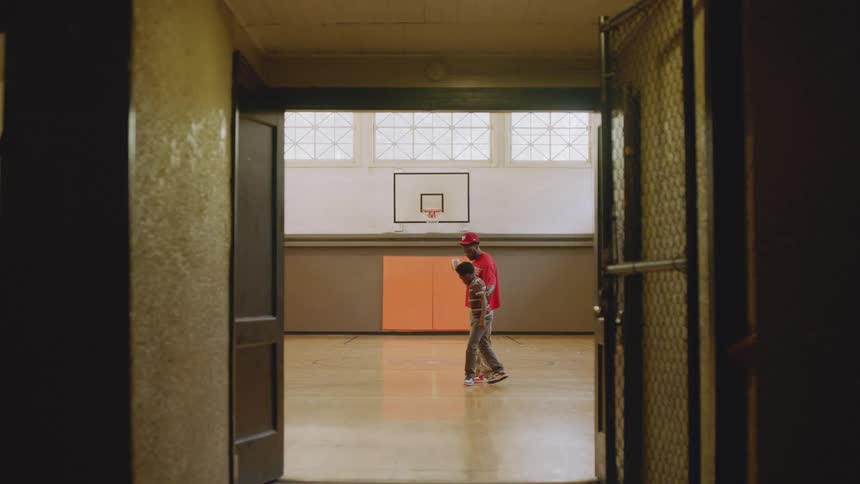 From a dark hallway, a father and his young son share a quiet, focused moment together in the middle of a brightly lit school gymnasium