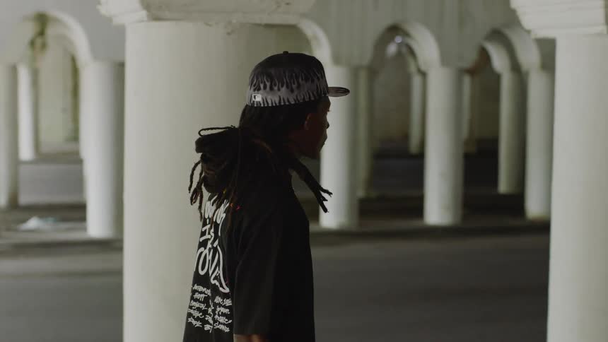 A man with dreadlocks stands in profile in an architectural underpass with repeating white columns as the camera slowly dollies right
