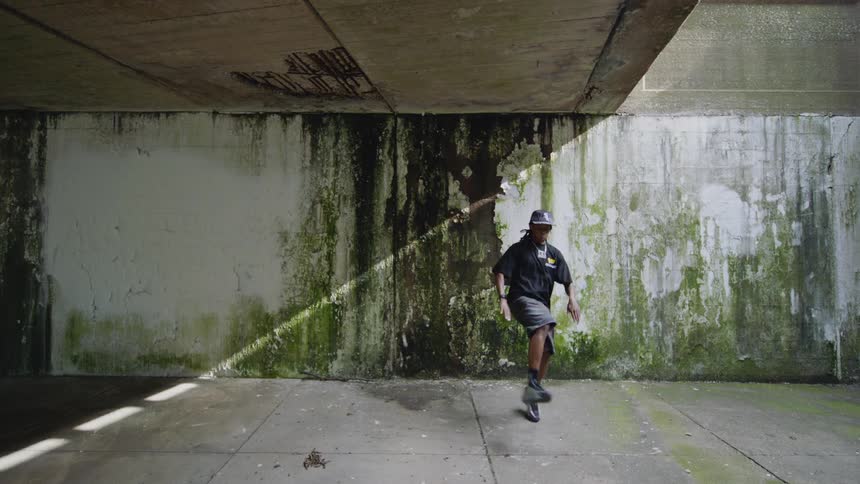 A young man performs an expressive hip-hop dance in slow motion under a gritty, moss-covered urban overpass