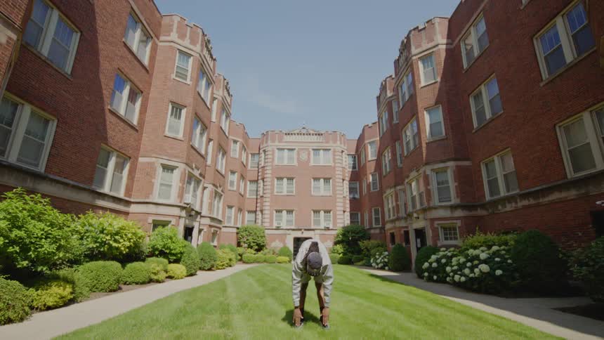 A young man in athletic wear warms up with stretches in the grassy courtyard of a large brick apartment building on a sunny day