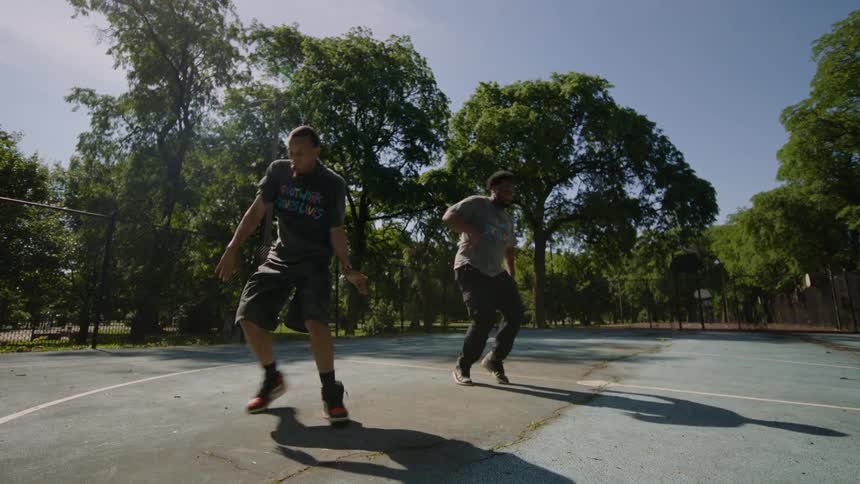 Slow-motion wide shot of two skilled male dancers performing a synchronized hip-hop routine on a sunny park basketball court