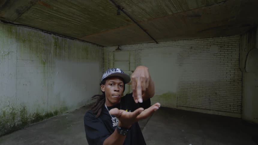 A confident young man with dreadlocks performs an intricate hand dance in a gritty, empty concrete room