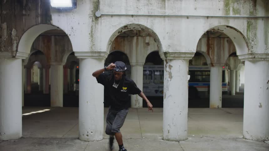 A young man performs expressive hip-hop dance moves in slow motion within a weathered, white-arched pavilion as a city bus passes by in the background