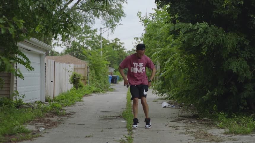 A young man showcases his intricate footwork while dancing down a gritty, overgrown urban alley