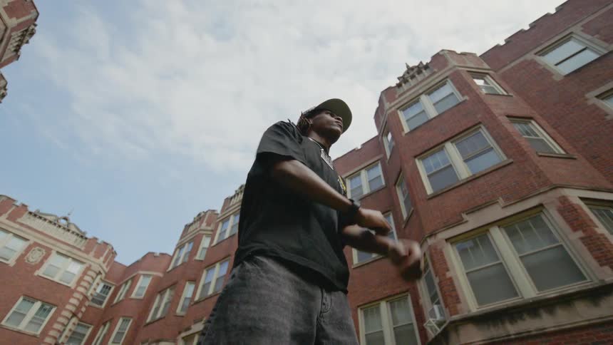 A young man with dreadlocks dances and stretches in slow motion in an urban courtyard, filmed from a low angle against brick apartment buildings