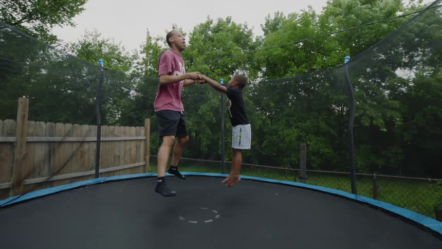 A loving father and his son hold hands while jumping together on a trampoline in their backyard in slow motion