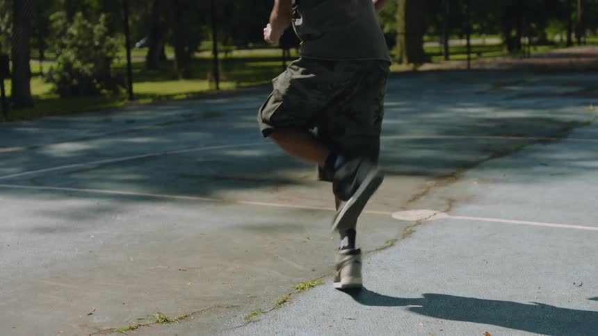 An energetic dancer with dreadlocks performs impressive footwork in slow motion on a basketball court in a sunlit park