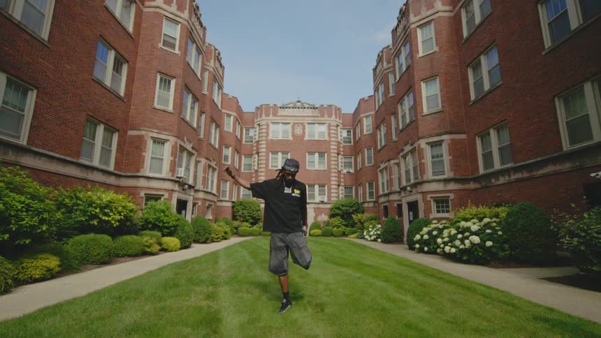 A young man with dreadlocks dances in slow motion in the grassy courtyard of a brick apartment complex