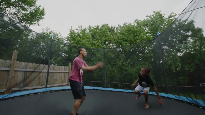 Slow motion footage shows a happy father and son jumping together on a trampoline in their backyard