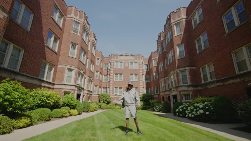 A young man in a grey sweatsuit performs a contemporary hip-hop dance in the grassy courtyard of a large brick apartment complex