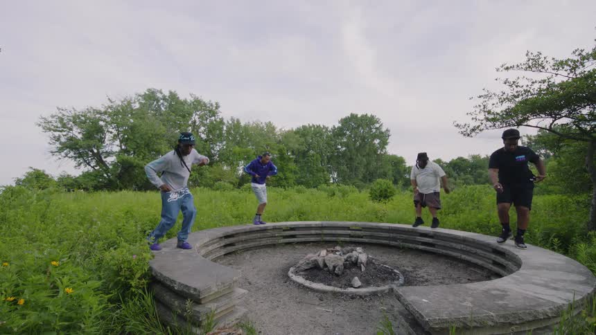 A group of four men perform an energetic, synchronized dance routine on a circular stone fire pit in a grassy park