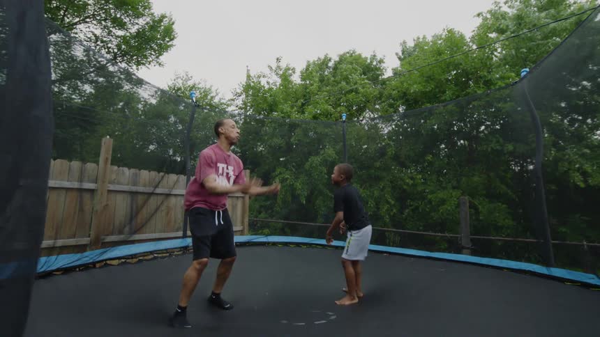 A father and son laugh and jump together in slow motion on a backyard trampoline