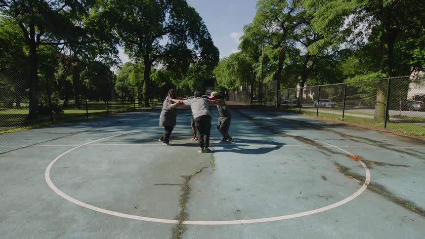 A diverse group of men performs an energetic and synchronized hip-hop dance routine on a sunny day at an urban basketball court
