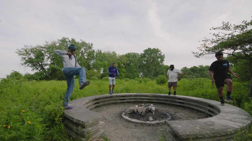 Four male dancers perform a synchronized routine in slow motion around a concrete fire pit in a grassy field